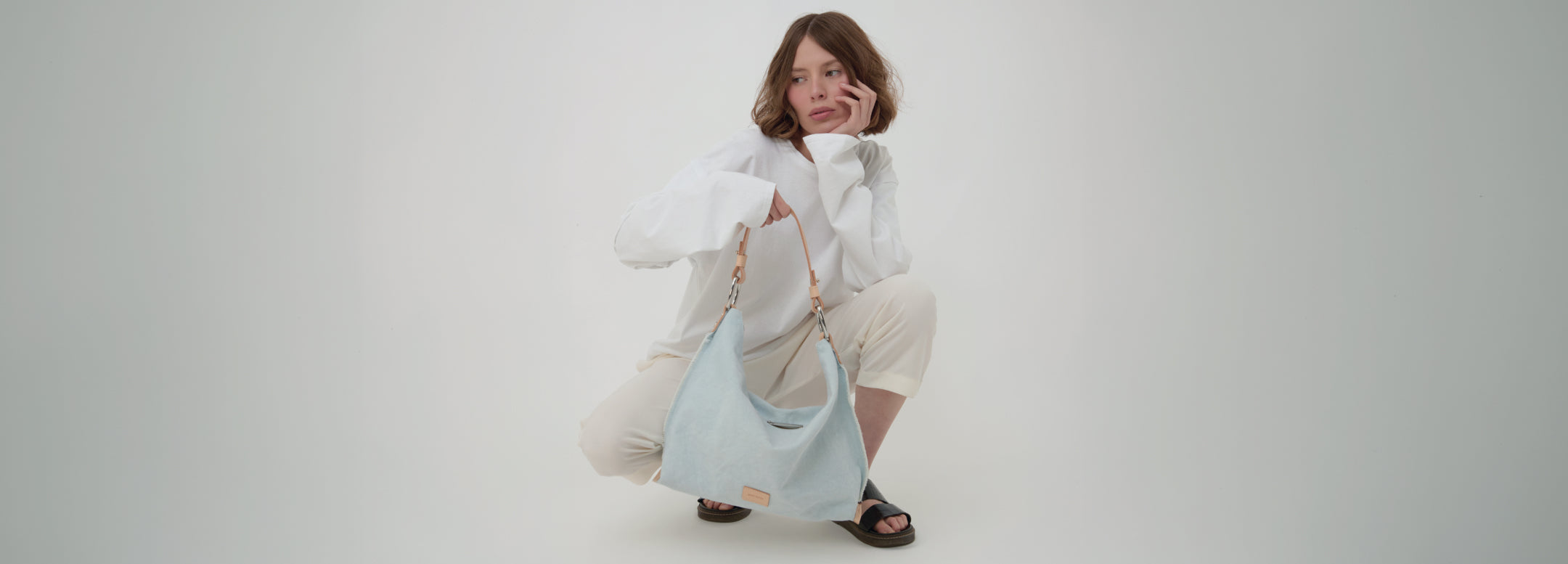 A person with long hair, wearing a denim jacket, is standing with their back to the camera. They are carrying a blue backpack with beige accents. Soft sunlight filters through nearby plants.