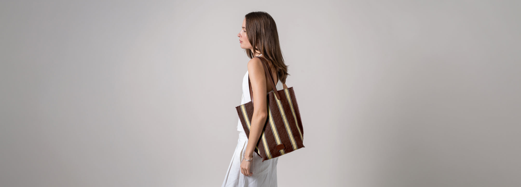 A person in a white shirt carries a brown felt laptop sleeve with a phone pocket. They are standing outdoors on a stone path with greenery and a door in the background.