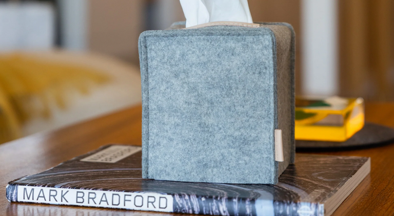 A grey felt tissue box sits on top of a book titled MARK BRADFORD, placed on a wooden table with blurred objects in the background.