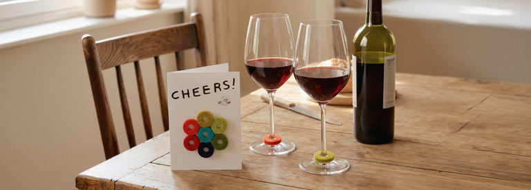 Two glasses of red wine and a bottle sit on a wooden table next to a chair. A greeting card reading CHEERS! and colorful wine glass markers are also on the table.