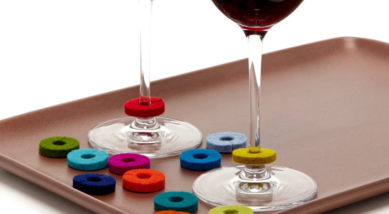 A cocktail glass with a green ring at the base stands on a white surface in front of a textured red background. A single cherry with a stem sits nearby.