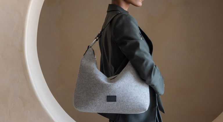 A person in a dark blazer carries a large, light gray shoulder bag with a black label, standing in front of a beige background with a circular design.