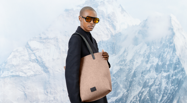 A person wearing a black coat and sunglasses holds a large bright red tote bag, standing in front of a snowy mountain backdrop.