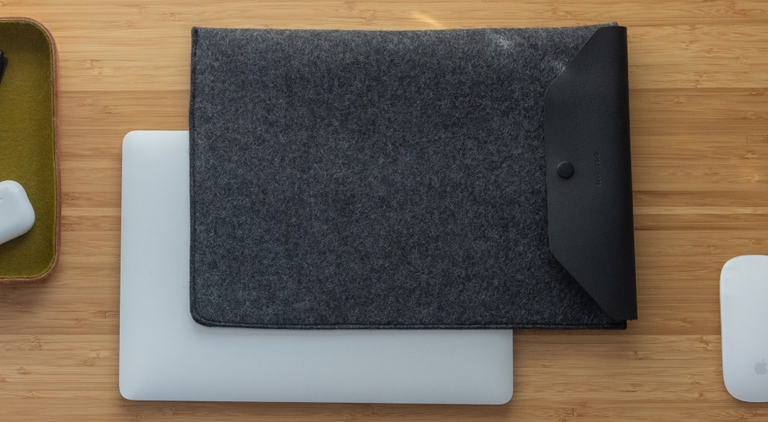 A laptop with a partially visible Apple logo sits on a scalloped mat on a wooden table. Nearby are a closed laptop sleeve and two pouches stacked on books. Soft shadows from tree branches dapple the scene.