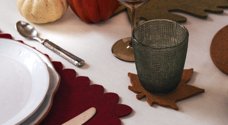 A glass of red beverage with a lemon slice on a pink coaster sits on a white slatted surface. Nearby are egg-shaped coasters in blue, green, and magenta.