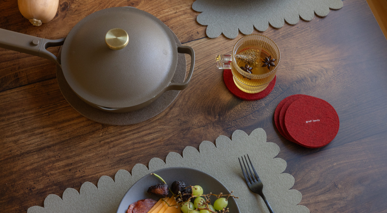 A person paints eggs at a table outdoors, surrounded by markers, a bowl of colorful eggs, fruit, lemons, and a drink. Bunny-shaped mats are on the table, adding a festive touch.