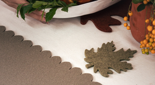 A wooden dining table set with blue placemats, black and wooden bowls of fruits and vegetables, a black pot, a glass mug, and a small flower vase. No people are present.