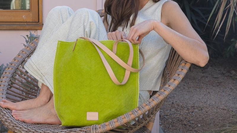 A woman in light, striped clothes sits barefoot on a wicker chair outdoors, holding an open lime green tote bag with tan handles on her lap.