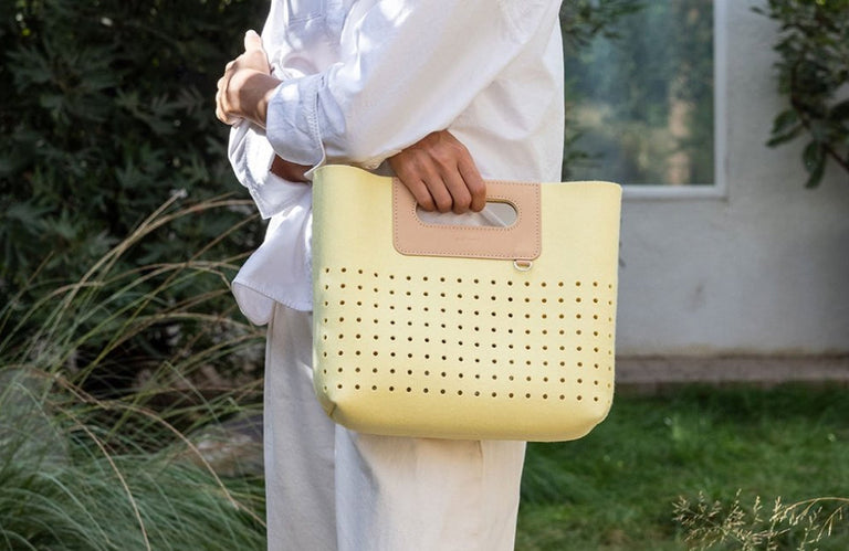 Person holding a pastel yellow perforated felt bag with a tan leather handle, standing outdoors in a lush garden setting. The stylish, modern tote is lightweight and perfect for spring and summer.