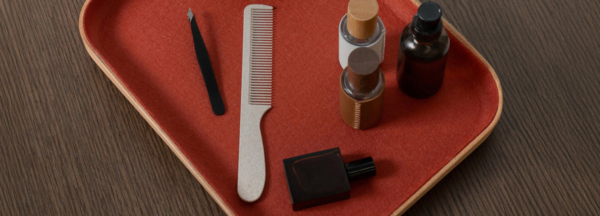 A red tray holds a white comb, black tweezers, two small bottles with wooden caps, a dark glass bottle, and a square perfume bottle on a dark wooden surface.