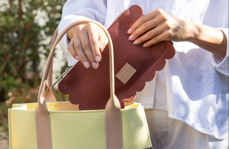 A person wearing a white shirt places a scalloped-edge, brown felt pouch into a yellow and beige tote bag outdoors.