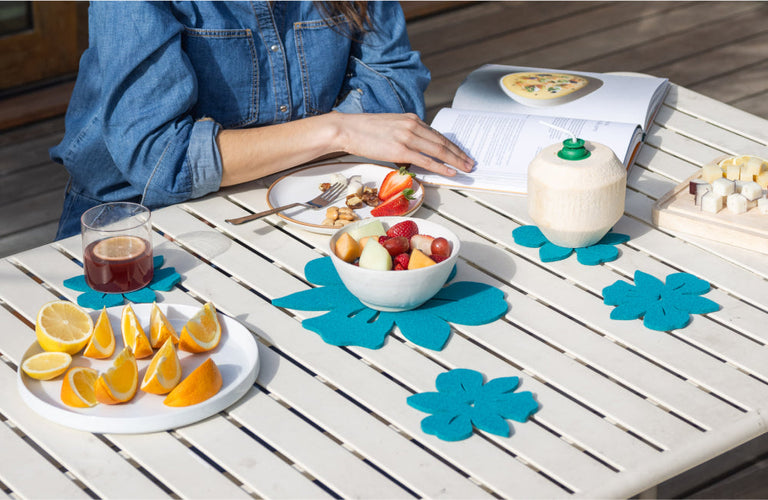 A person in a denim shirt sits at a table outdoors with fruit, a drink, a coconut, and an open cookbook. Blue flower-shaped placemats decorate the white table.