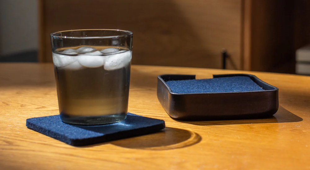 A glass of iced water sits on a dark blue coaster on a wooden table, with a matching coaster holder placed nearby. Warm sunlight illuminates the scene.