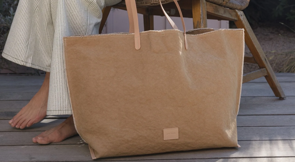 A large tan tote bag with leather handles sits on a wooden deck next to a person wearing light-colored, striped pants, with bare feet visible.