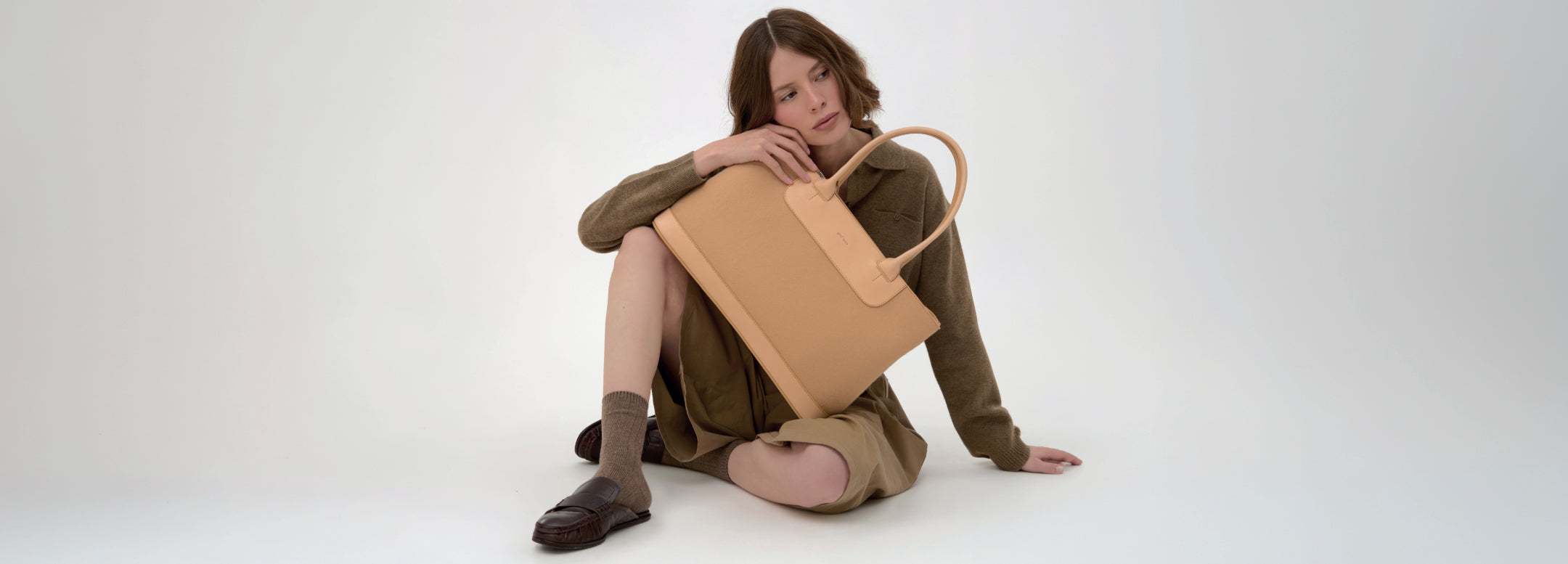 A person wearing a cream sweater and beige pants holds a large olive-green tote bag with tan leather handles and accents against a plain light background.