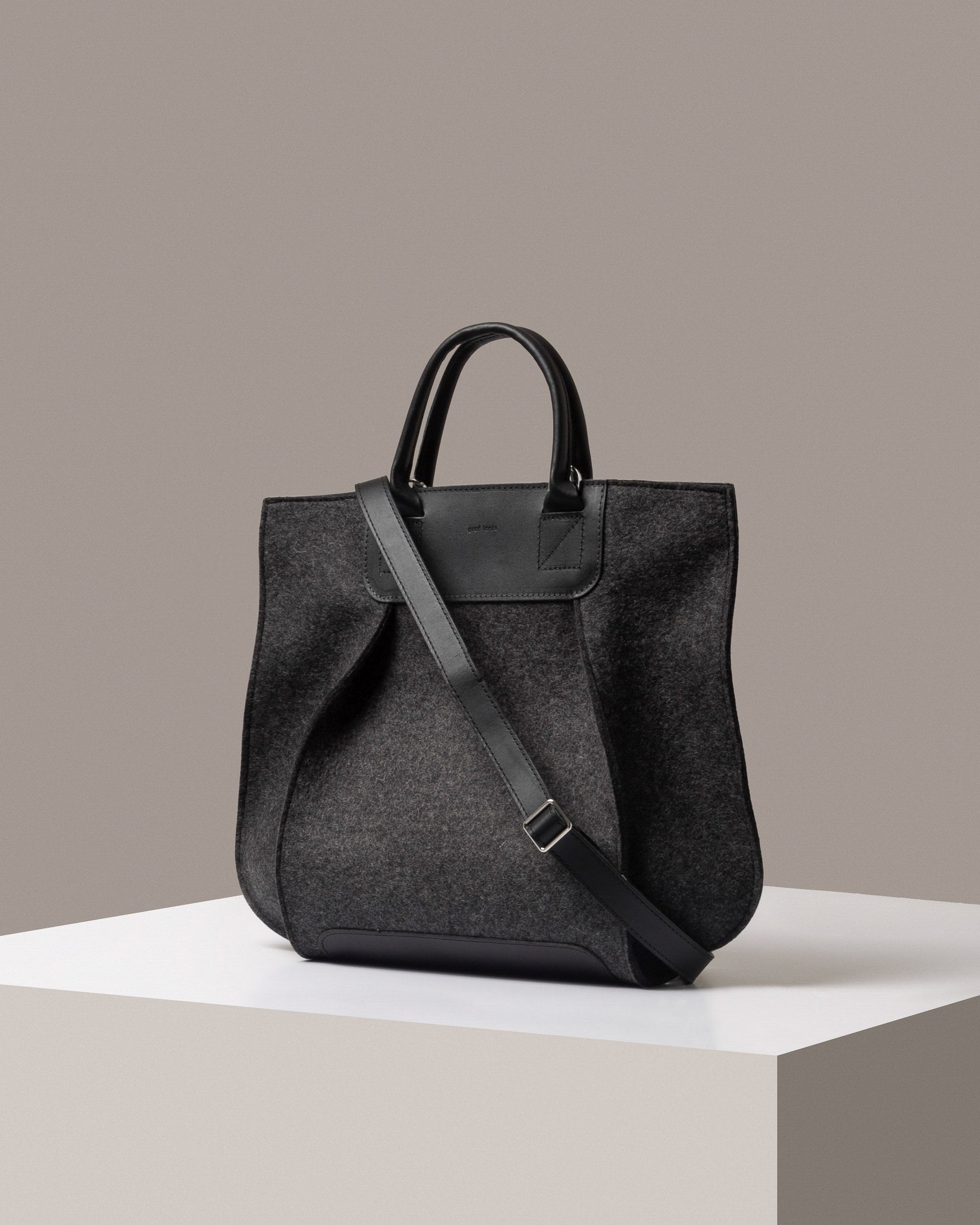 A stylish Frankie Merino Wool Felt Tote bag in dark gray with a black leather shoulder strap, displayed in a three-quarter view on a white base