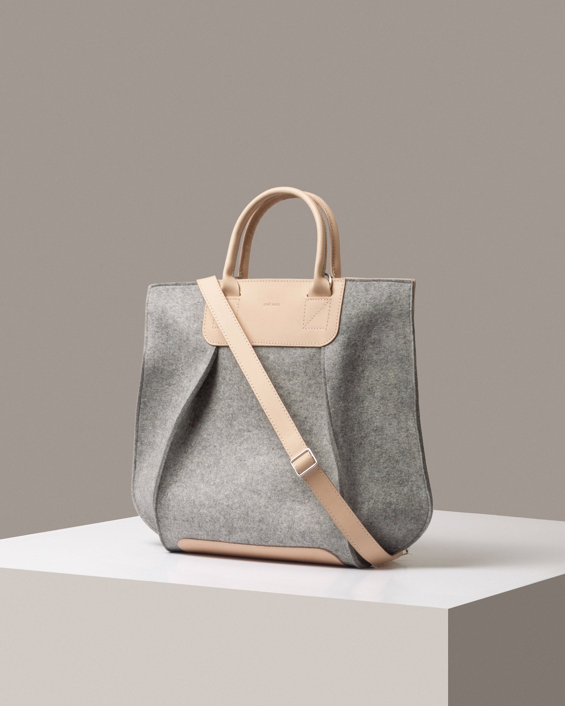 A stylish Frankie Merino Wool Felt Tote and crossbody bag in gray with a beige leather shoulder strap, displayed in a three-quarter view on a white base