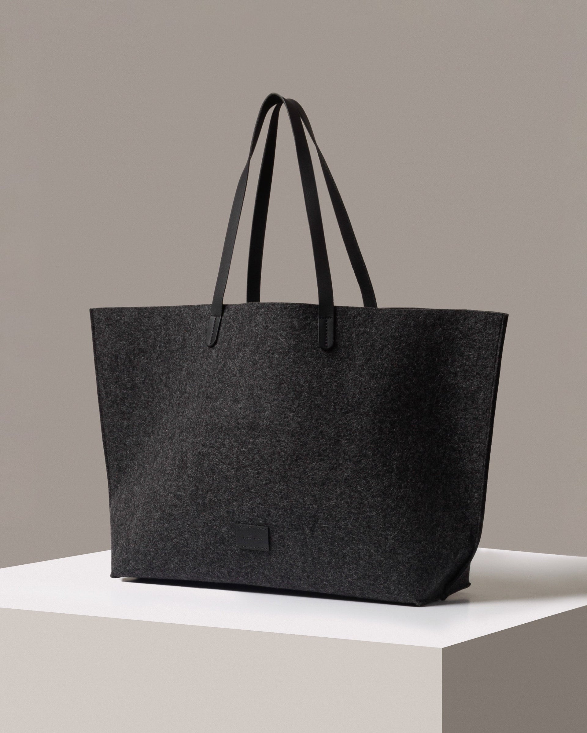 A stylish Hana Merino Wool Felt Boat Bag in dark gray with black leather handles, displayed in a three-quarter view on a white base