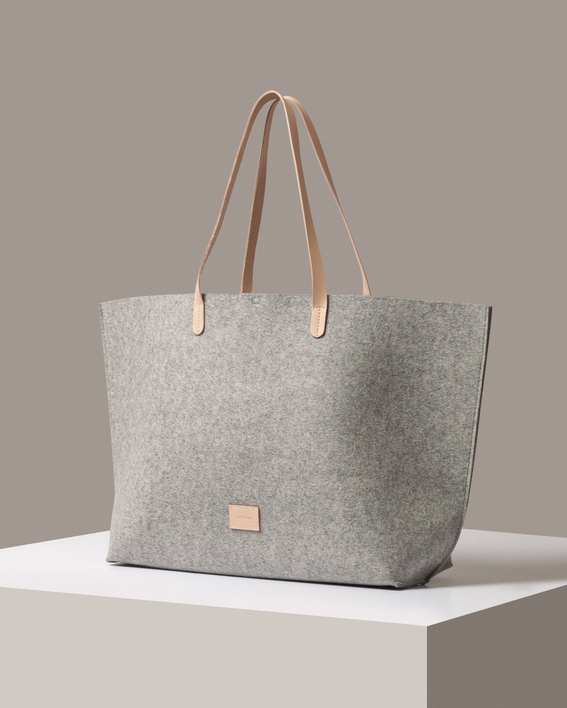 A stylish Hana Merino Wool Felt Boat Bag in gray with beige leather handles, displayed in a three-quarter view on a white base