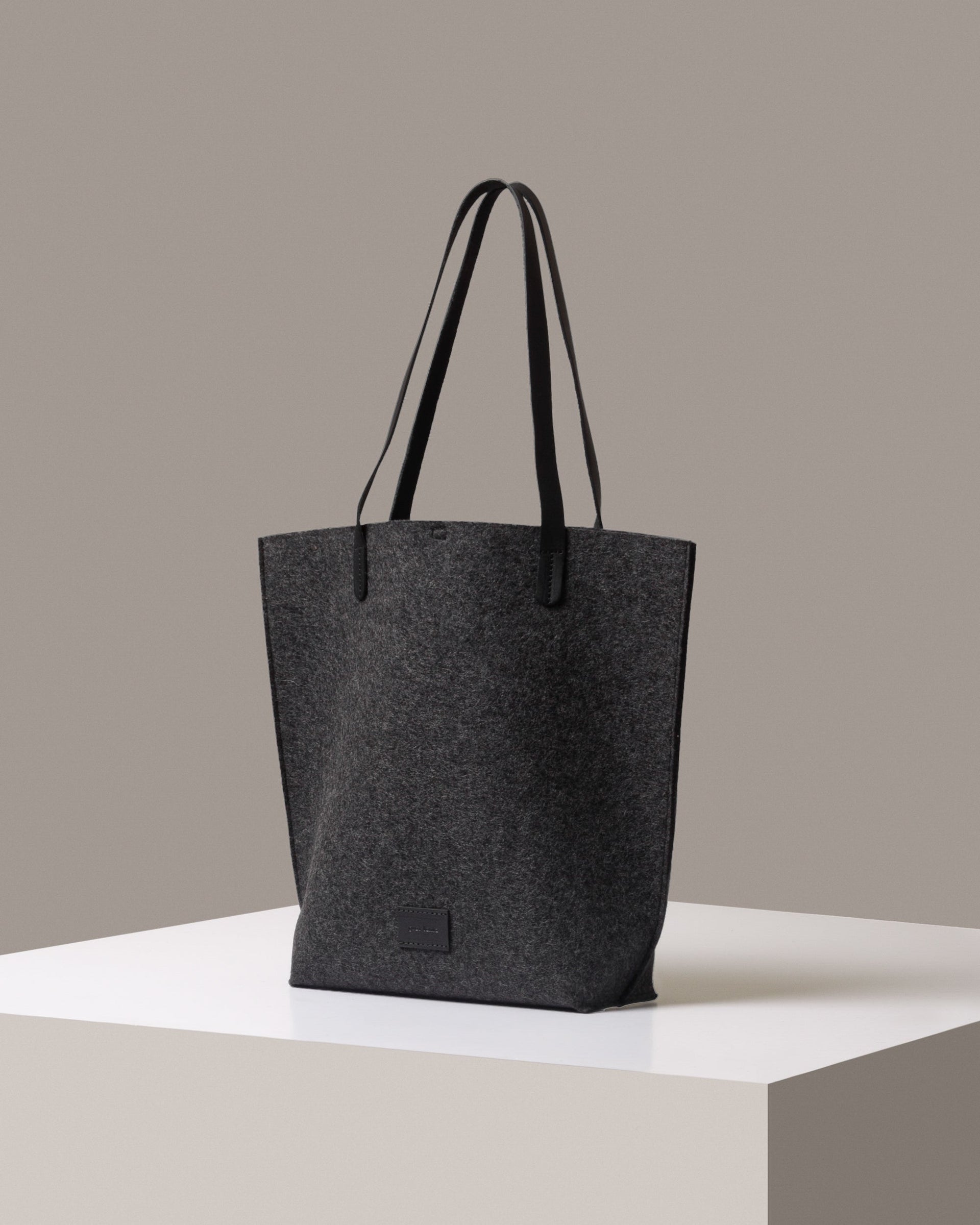 A Hana Merino Wool Felt Tote bag in dark gray with black leather handles, displayed in a three-quarter view on a white base