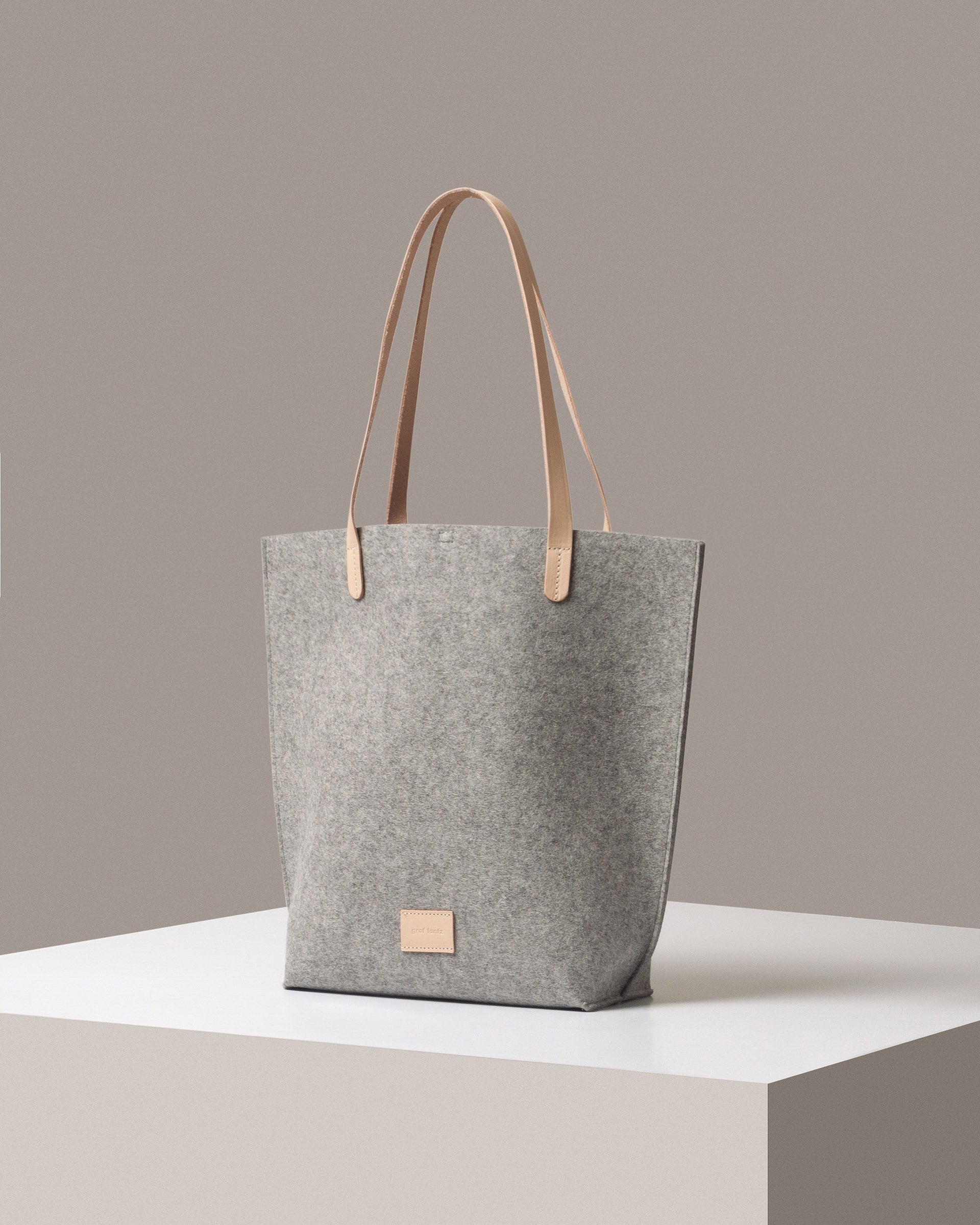 A Hana Merino Wool Felt Tote bag in gray with beige leather handles, displayed in a three-quarter view on a white base