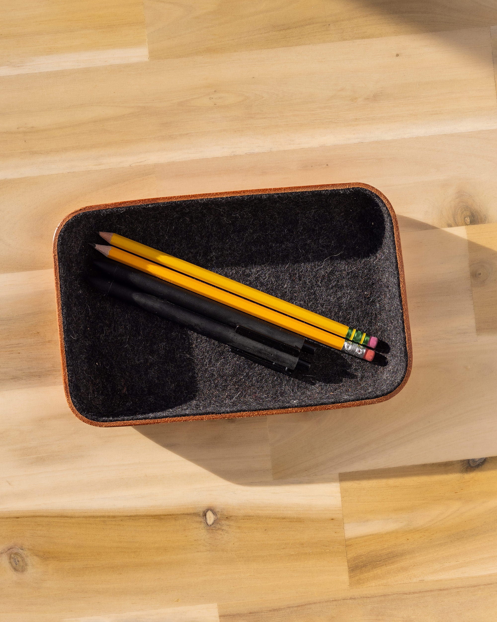 A rectangular catchall tray in Vachetta leather by graf lantz rests on a wooden surface, holding two yellow pencils and a black pen. Shadows elegantly enhance the warm, minimalistic ambiance of the scene.