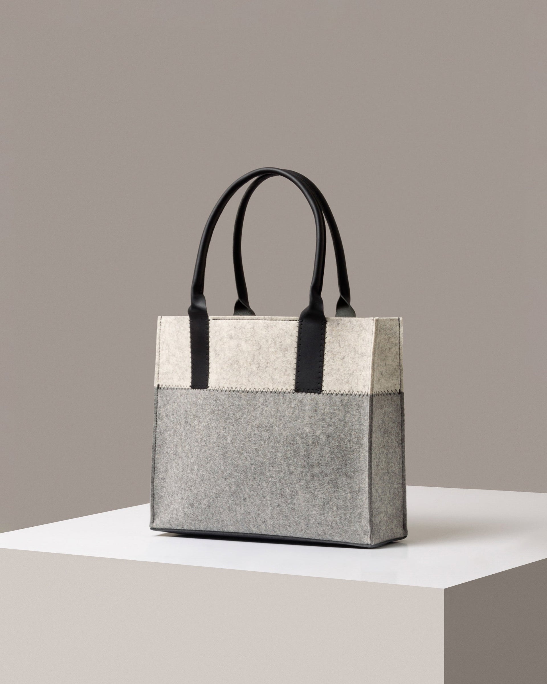 A stylish Jaunt Midi Merino Wool Felt Tote bag in white, gray, and black colors, displayed in a three-quarter view on a white base