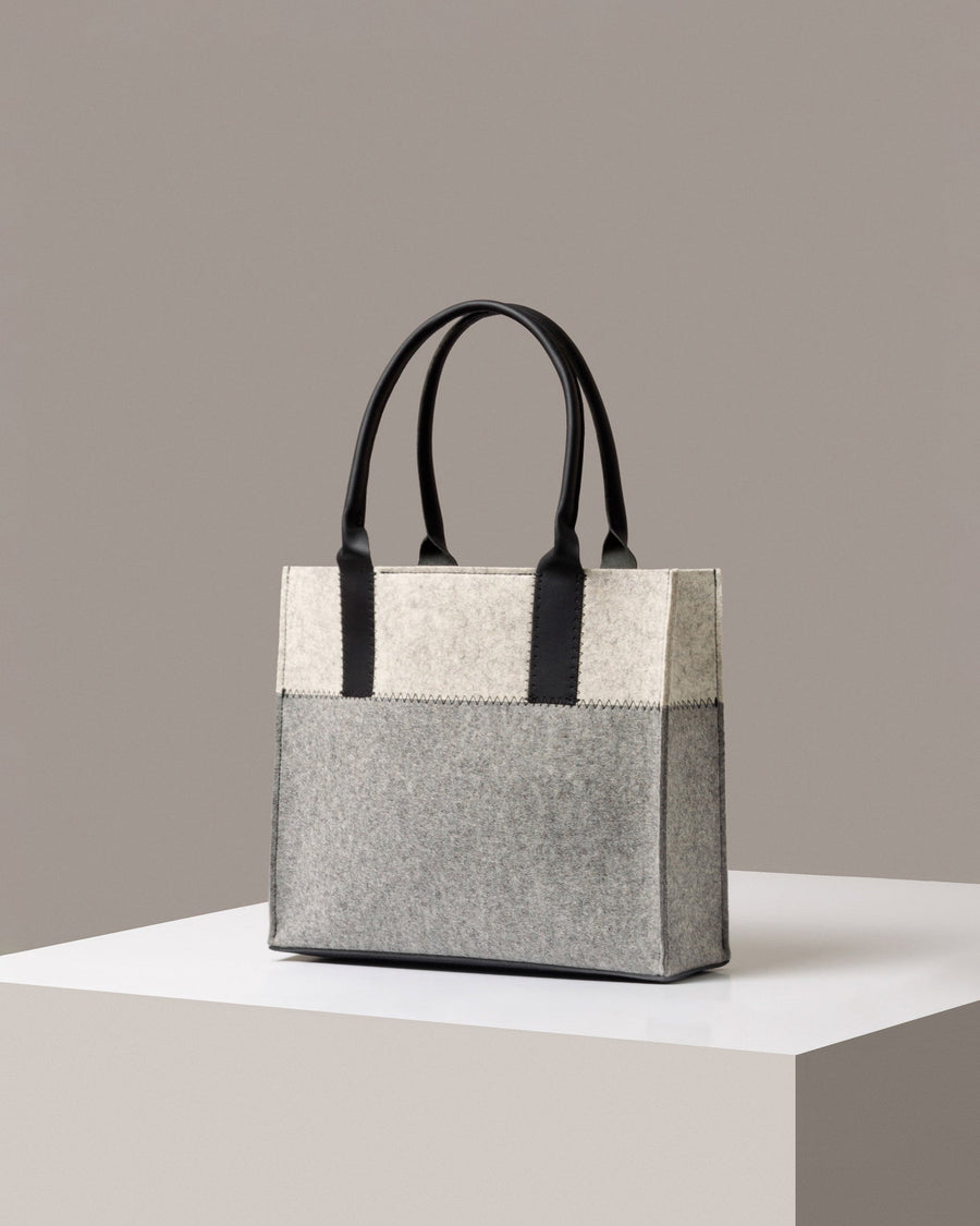 A stylish Jaunt Midi Merino Wool Felt Tote bag in white, gray, and black colors, displayed in a three-quarter view on a white base
