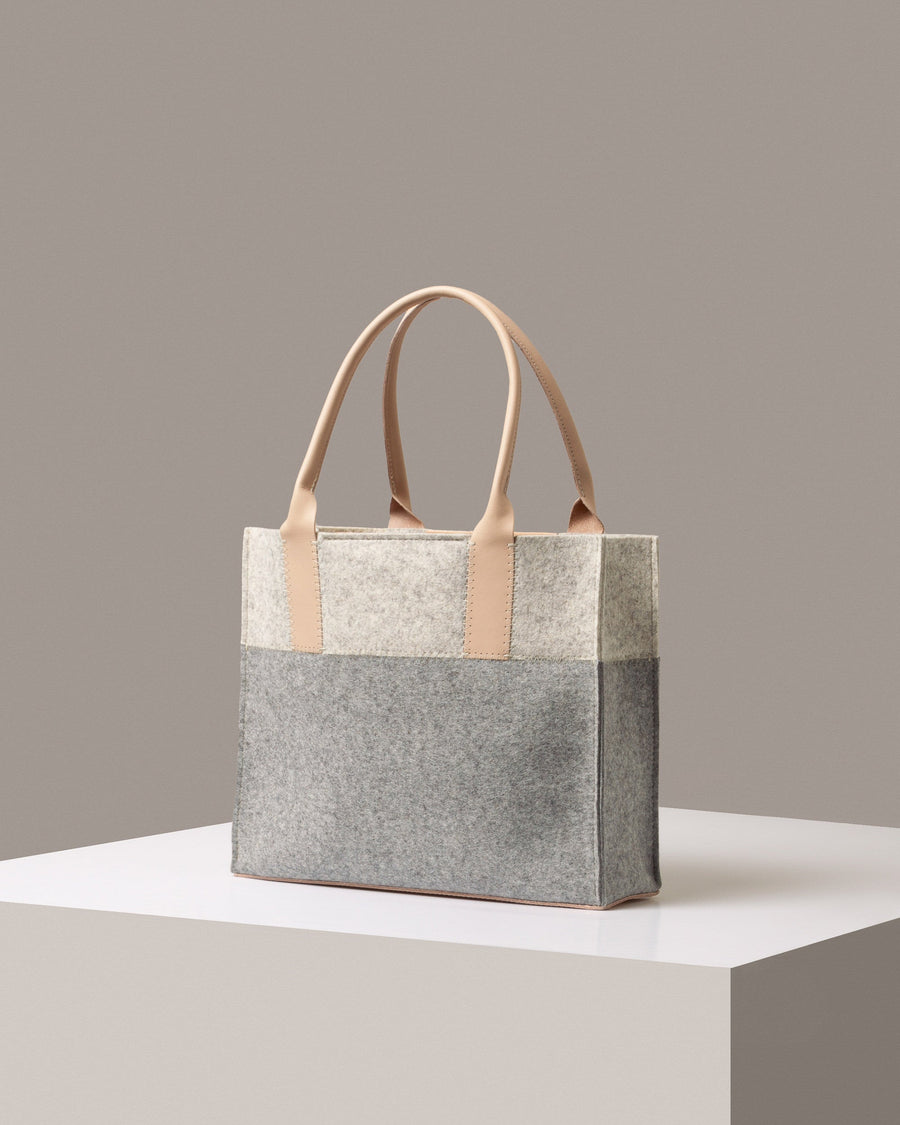 A stylish Jaunt Midi Merino Wool Felt Tote bag in white, gray, and beige colors, displayed in a three-quarter view on a white base