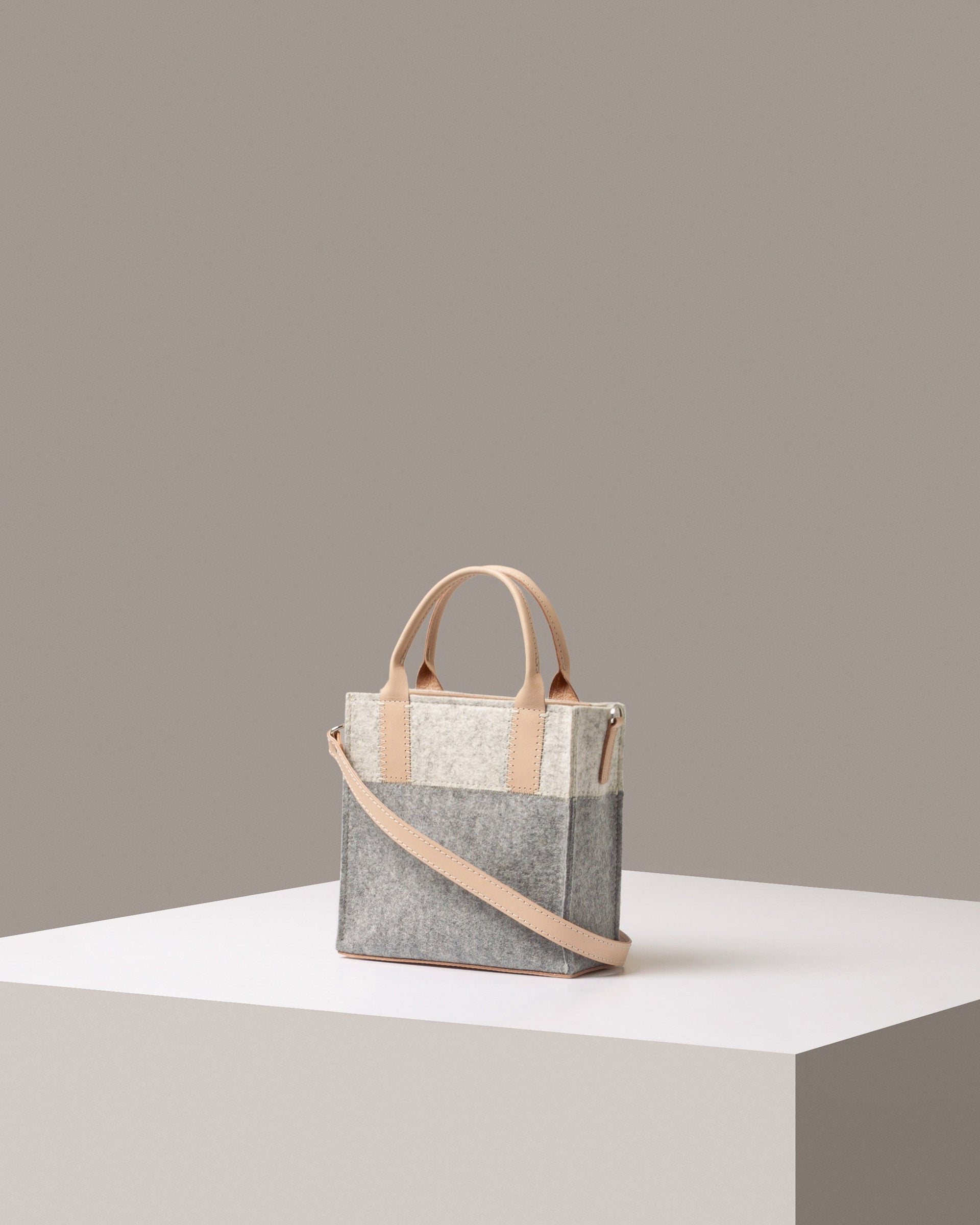 A stylish Jaunt Mini Merino Wool Felt Crossbody bag in white, gray, and beige colors, displayed in a three-quarter view on a white base