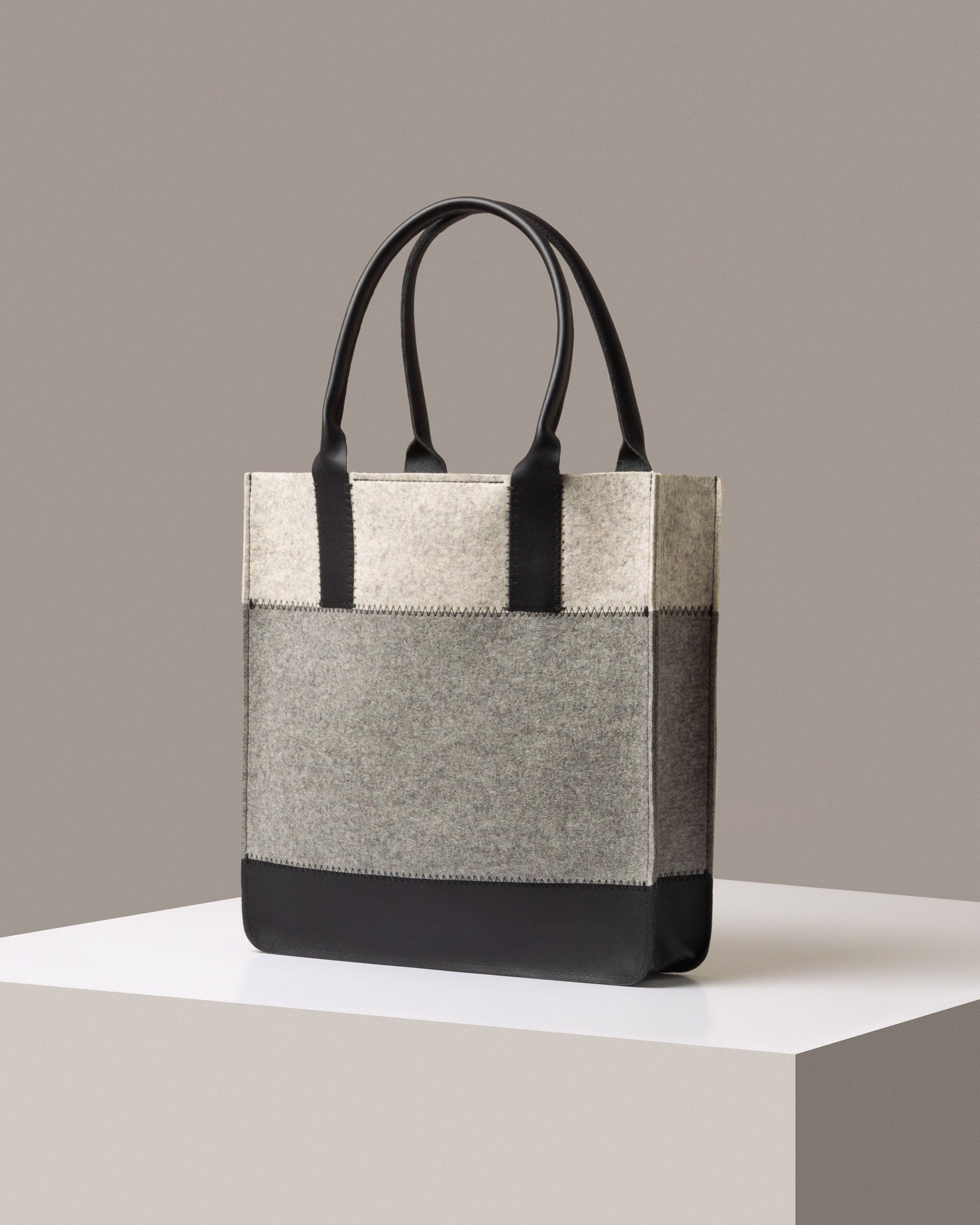 A stylish Jaunt Merino Wool Felt Tote bag in gray, white, and black colors, displayed in a three-quarter view on a white base