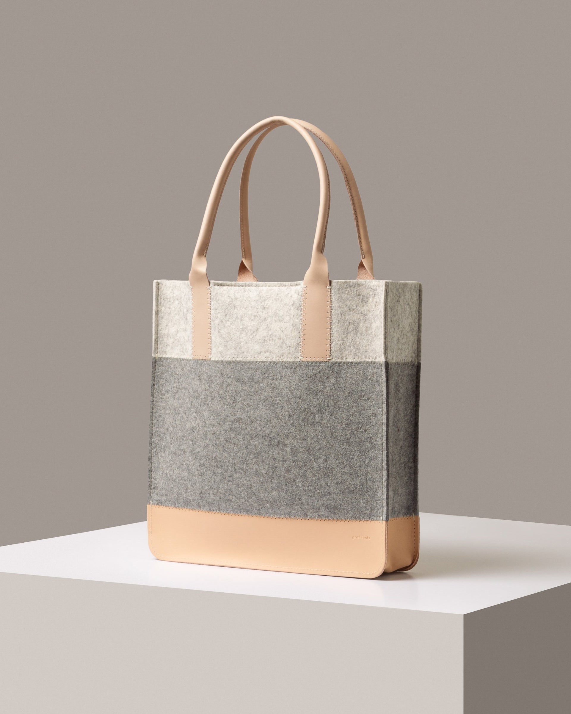 A stylish Jaunt Merino Wool Felt Tote bag in white, gray, and beige colors, displayed in a three-quarter view on a white base