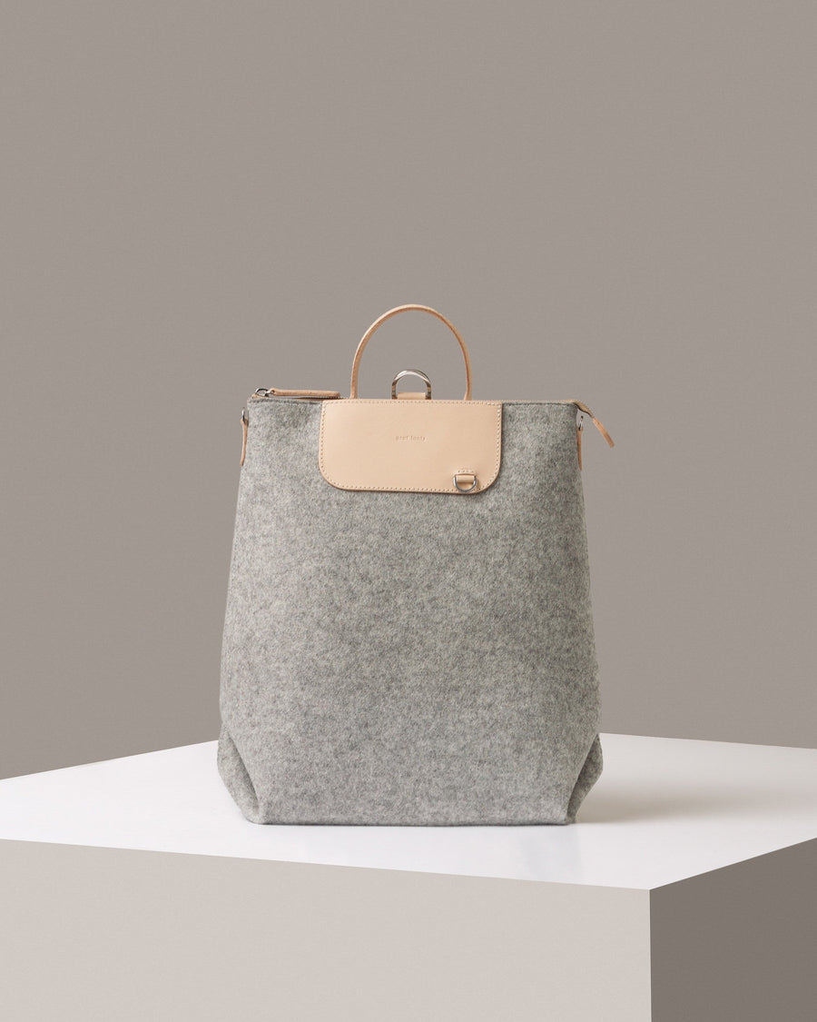 A Bedford Merino Wool Felt Backpack in light grey with beige leather accents standing on a white base, front view