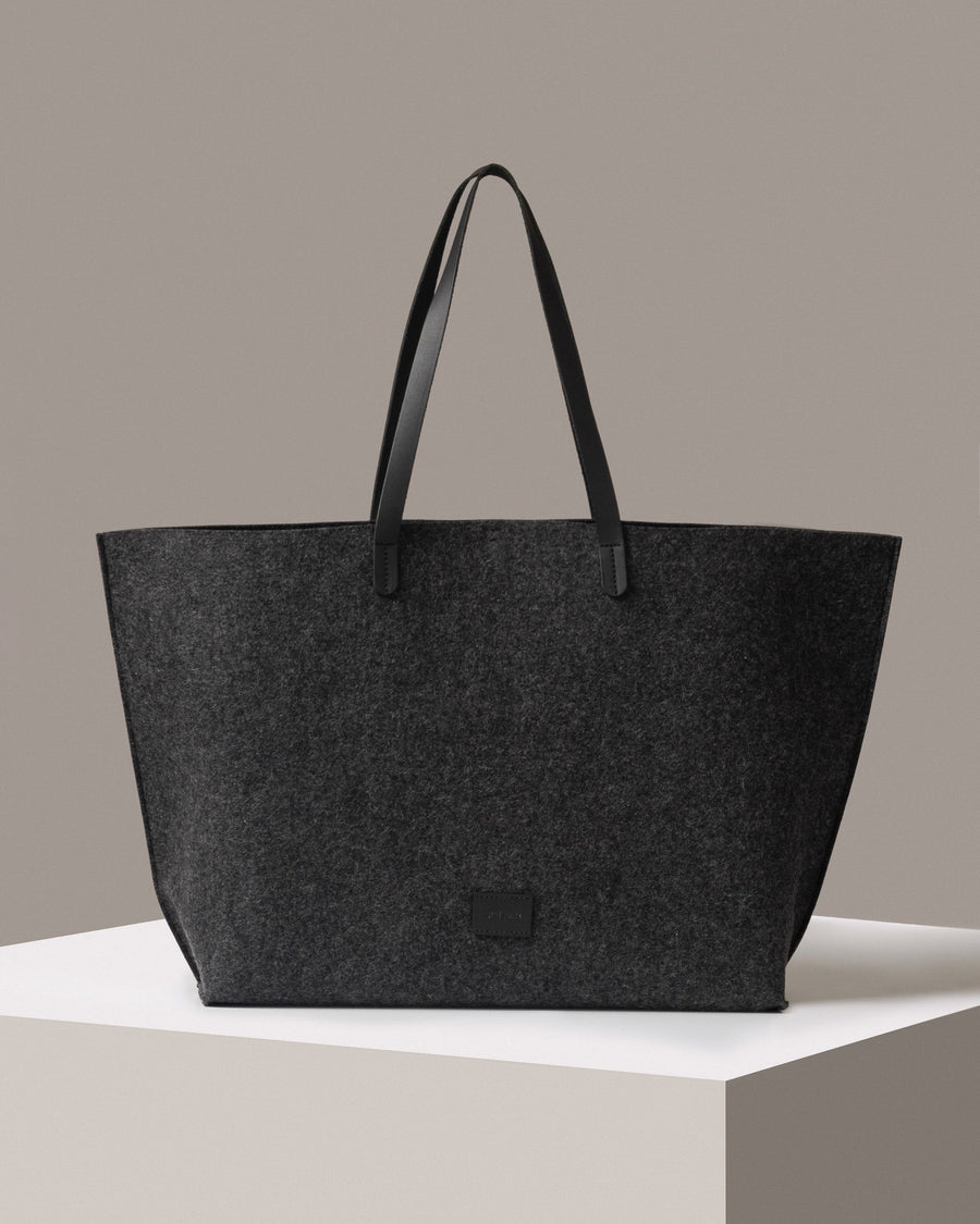 A Hana Merino Wool Felt Boat Bag in dark grey with black handles standing on a white base, front view