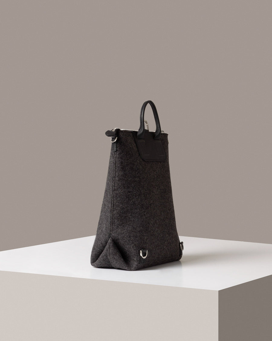 A dark grey Bedford Merino Wool Felt Backpack on a white base displayed in a side-view
