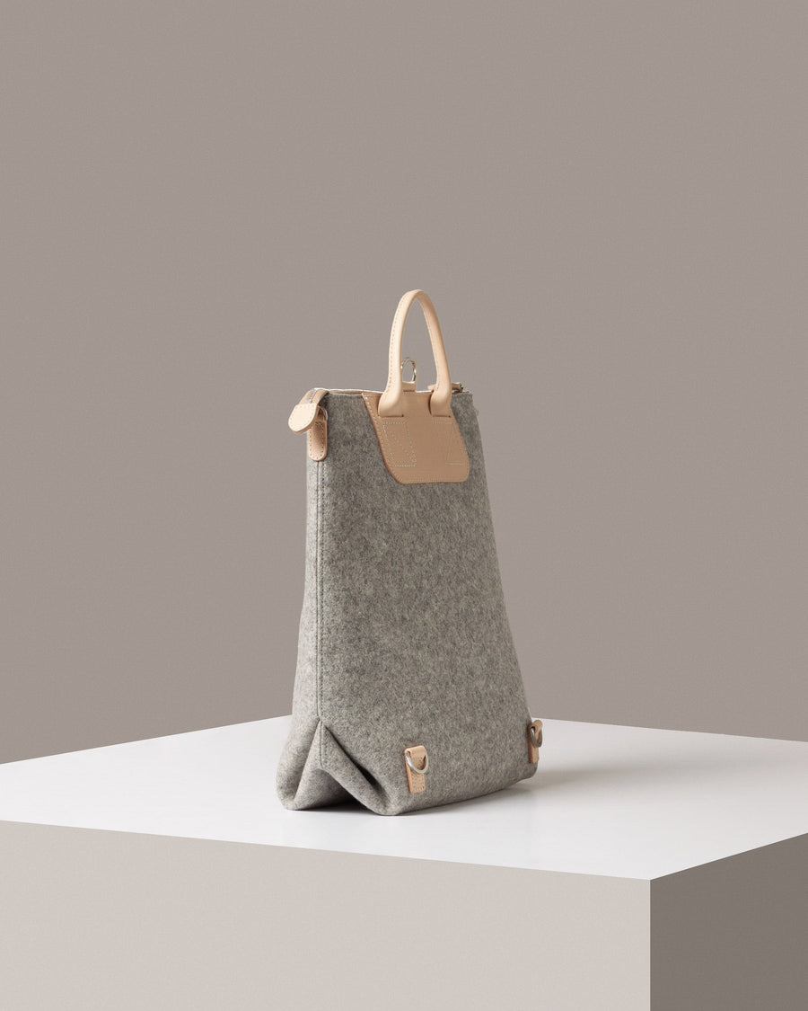 A grey Bedford Merino Wool Felt Backpack on a white base is displayed in a side-view
