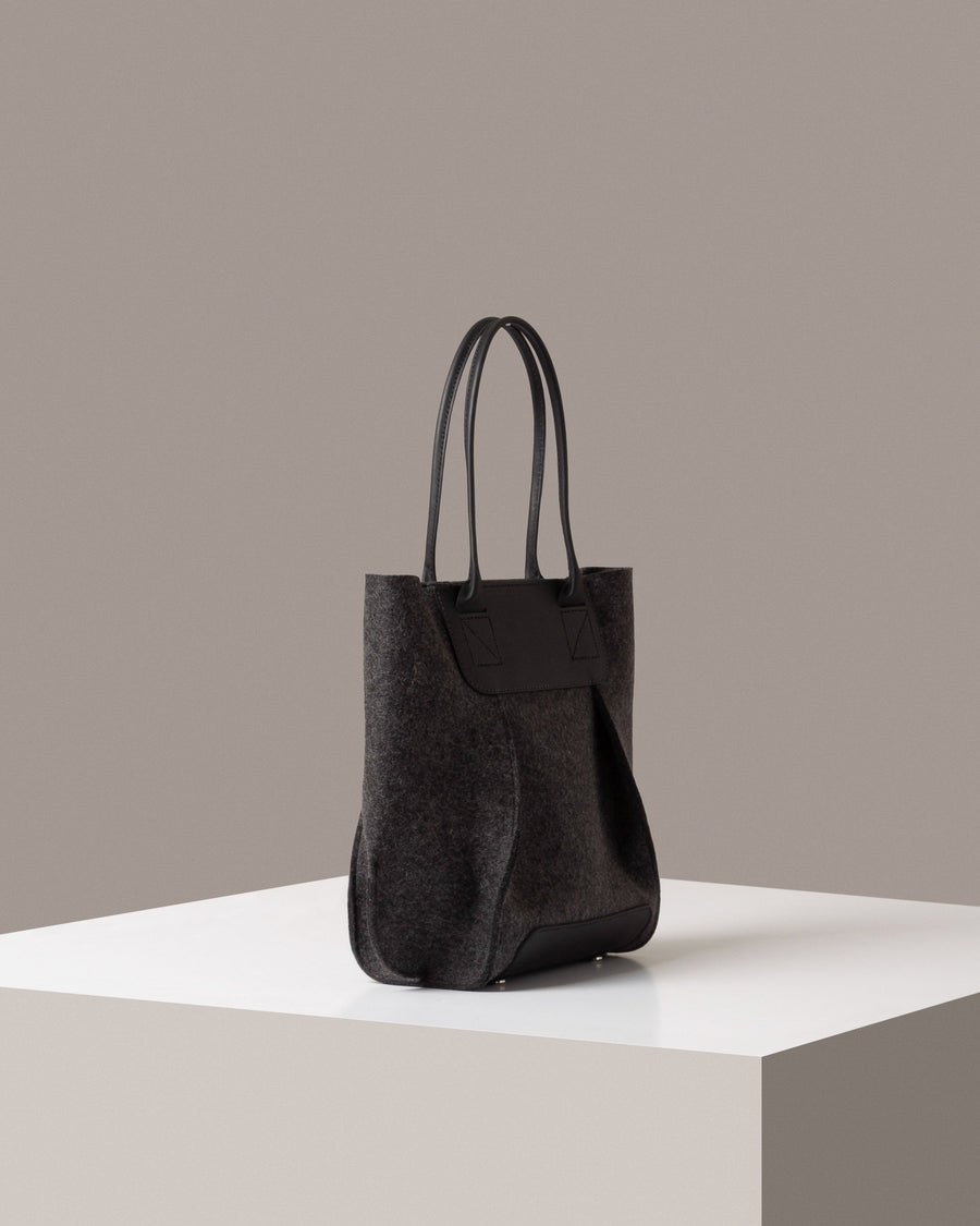 A dark grey Frankie Merino Wool Felt Midi mid-sized tote bag on a white base displayed in a side-view
