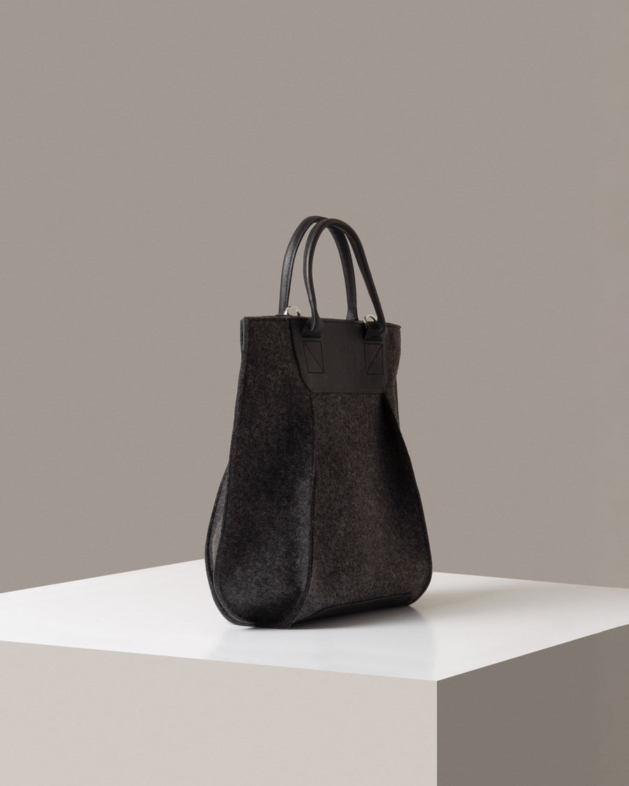 A dark grey Frankie Merino Wool Felt Tote on a white base displayed in a side-view
