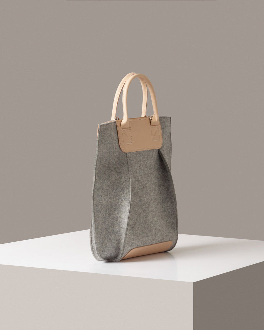 A Frankie Merino Wool Felt Tote in a grey color with beige leather accents on a white base displayed in a side-view
