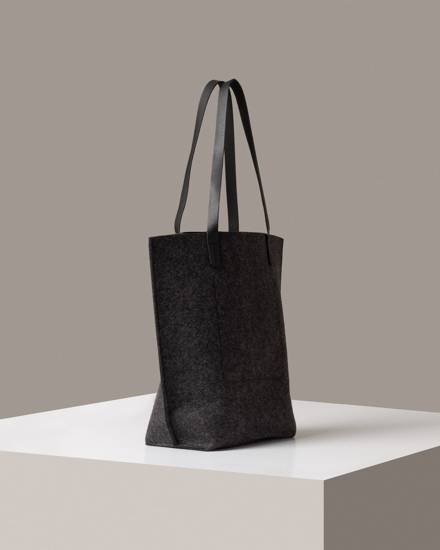 A dark grey felt tote bag with black leather handles on a white base displayed in a side-view

