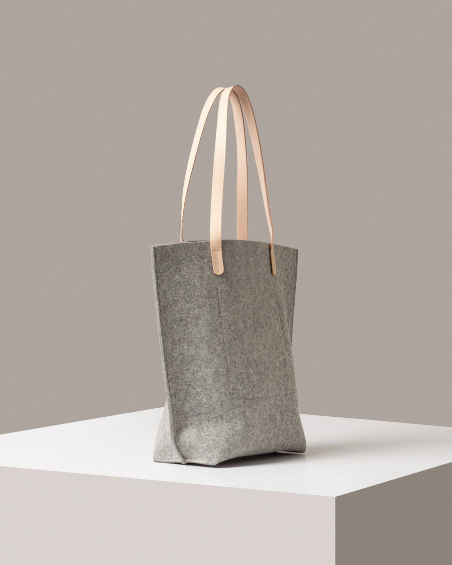 A grey felt tote bag with beige leather handles on a white base displayed in a side-view
