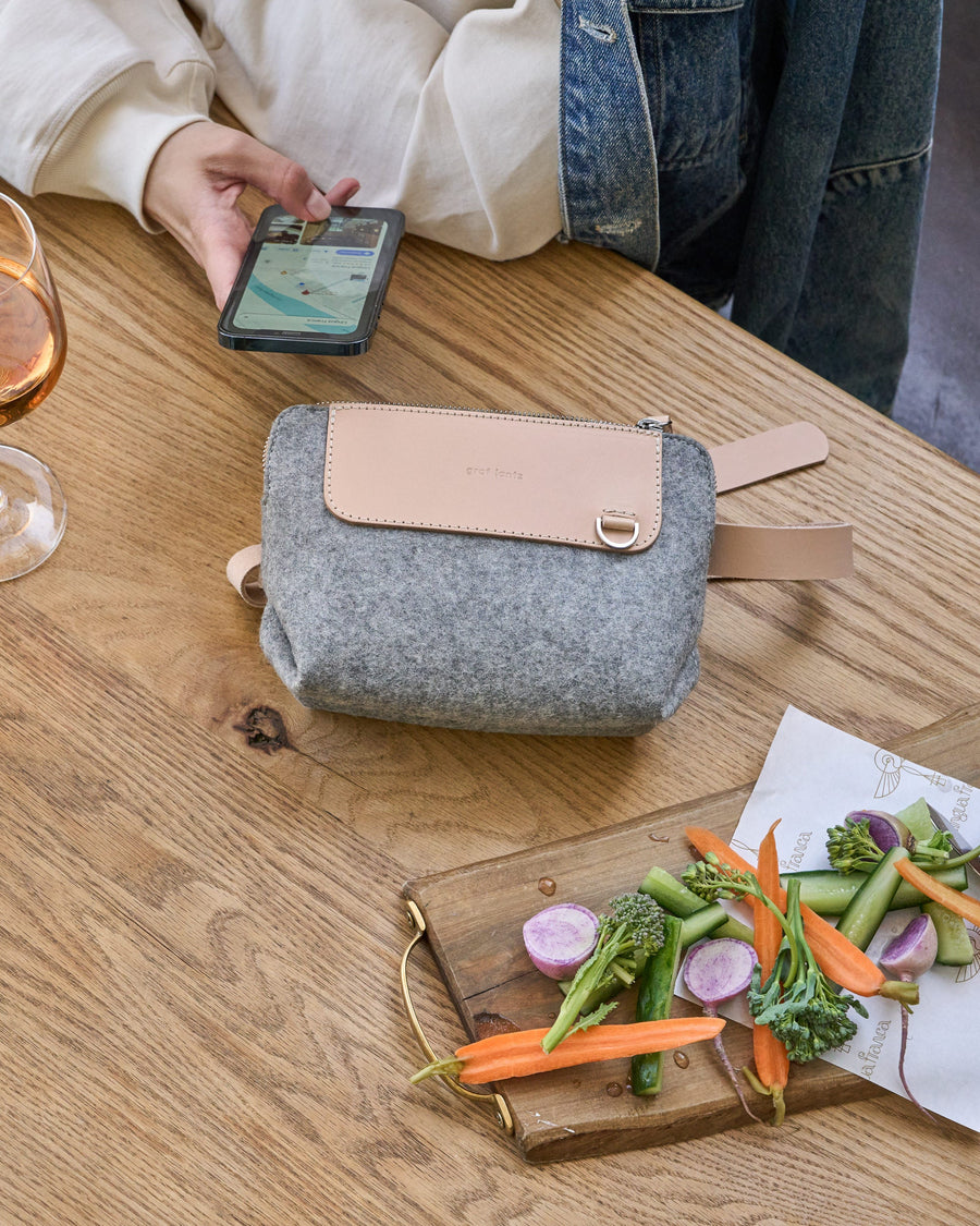 A granite-colored Bedford Merino Wool Felt Belt Bag placed on a wooden table beside a wine glass and a plate with vegetable finger food