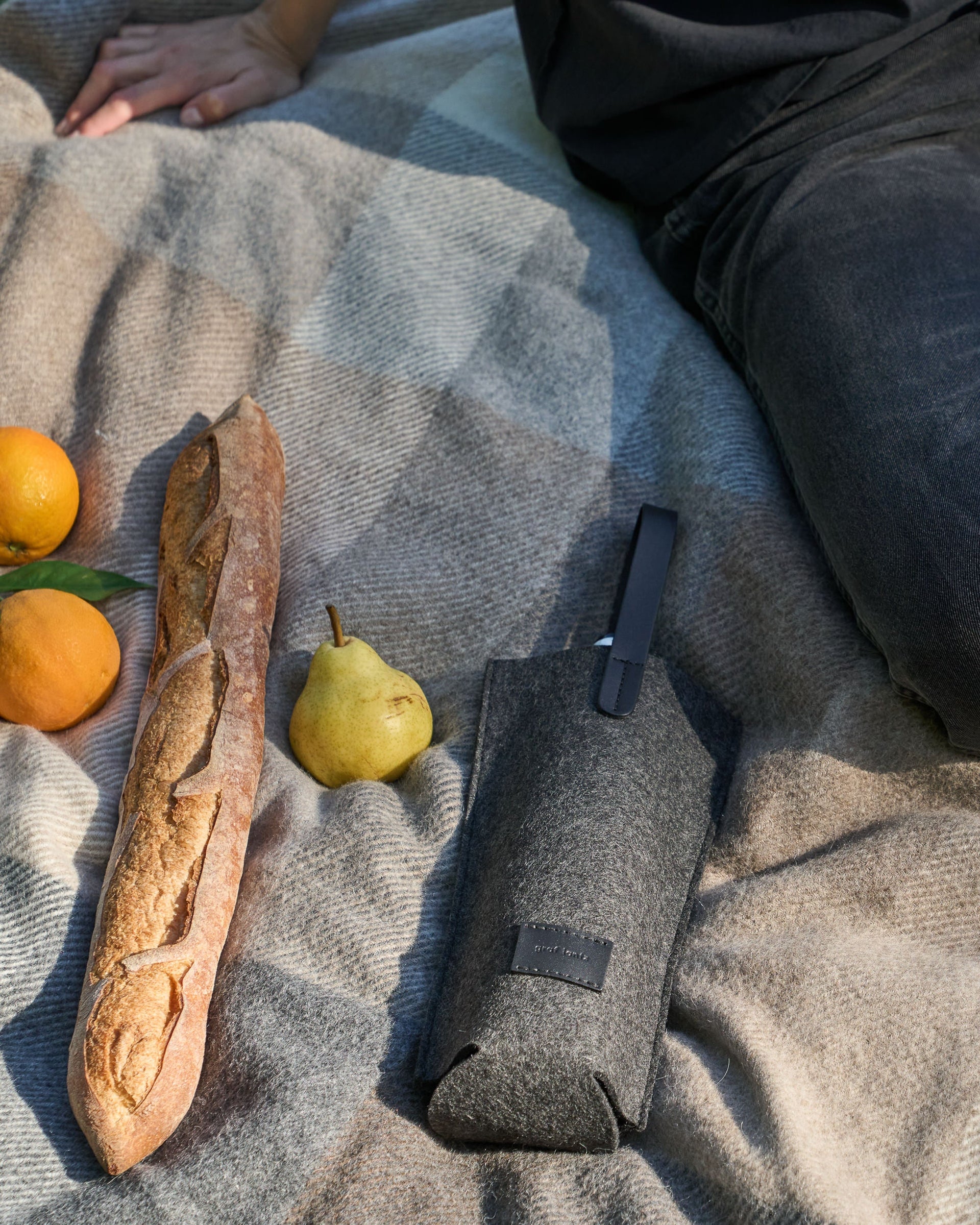 A charcoal-colored Hana Merino Wool Bottle Bag with black leather handle, styled on a picnic blanket with fruits and a baguette
