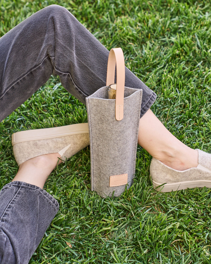 Granite-colored Hana Merino Wool Bottle Bag with beige leather handle, featuring bottle inside, set on lush green grass beside seated person