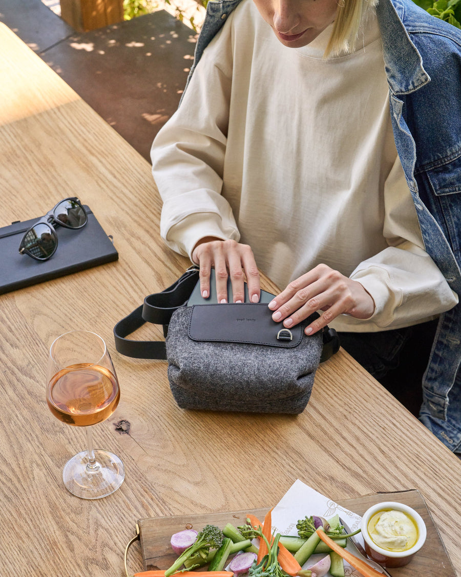 Hands retrieving a wallet from a charcoal-colored Bedford Merino Wool Felt Belt Bag placed on a wooden table beside a wine glass and a plate of vegetables