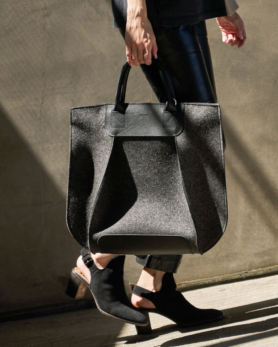 A charcoal-colored Frankie Merino Wool Felt Tote held by the top handle on a woman's side while walking, highlighting its chic design
