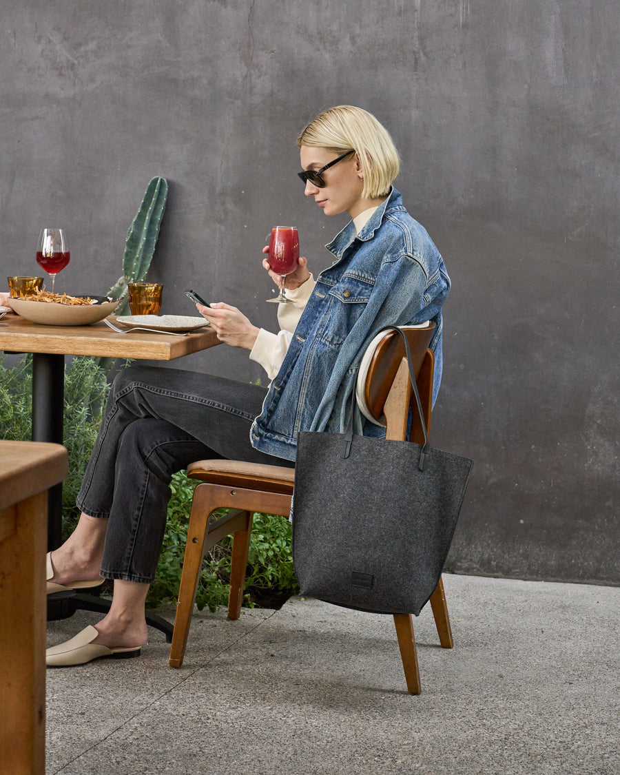 Chic woman sitting in a restaurant with the leather straps of a Hana Merino Wool Felt Tote elegantly hanging over the chair back