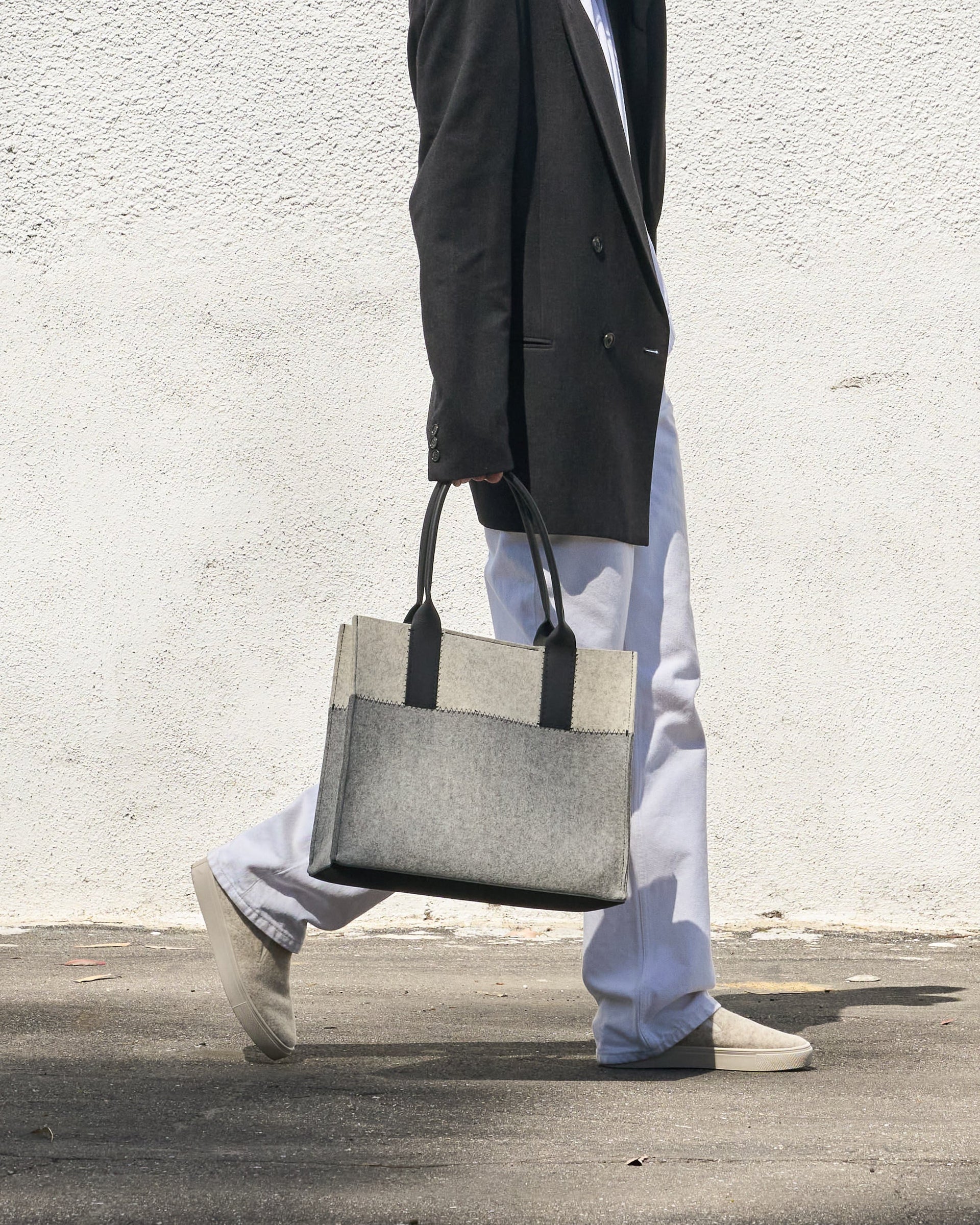 A grey, white, and black colored Jaunt Midi Merino Wool Felt Tote bag is elegantly held by its top handle as a woman walks, showcasing its chic design