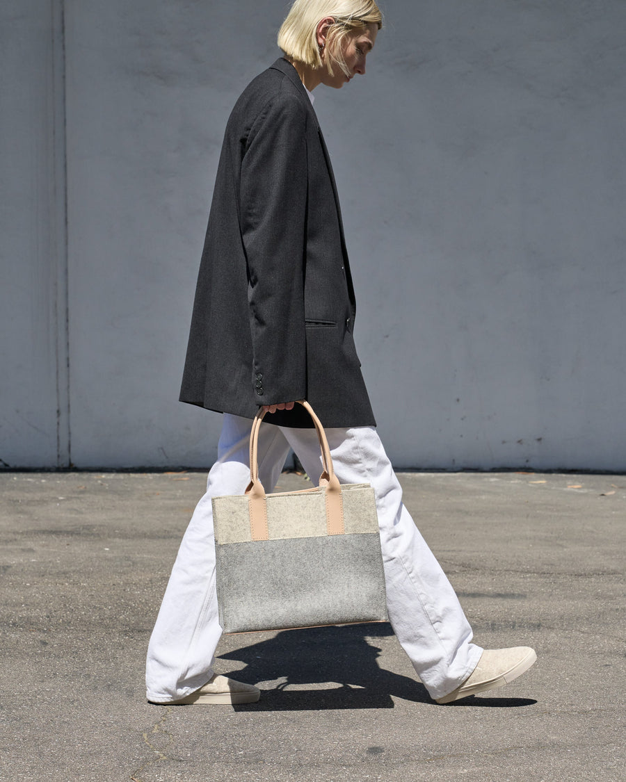 A grey, white, and beige colored Jaunt Midi Merino Wool Felt Tote bag is elegantly held by its top handle as a woman walks, showcasing its chic design