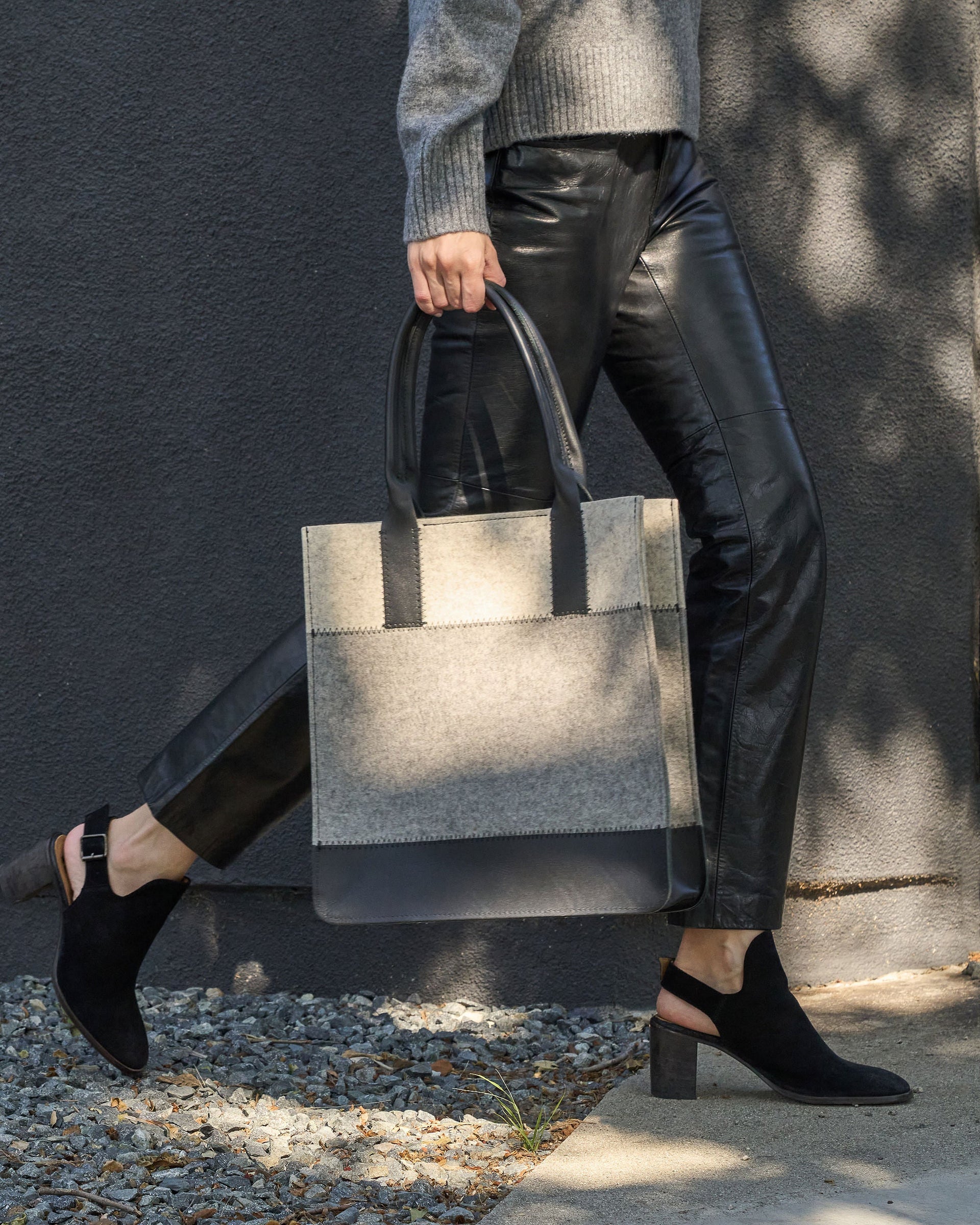 A dark grey, white, and black colored Jaunt Merino Wool Felt Tote bag is elegantly held by its top handle as a woman walks, showcasing its chic design